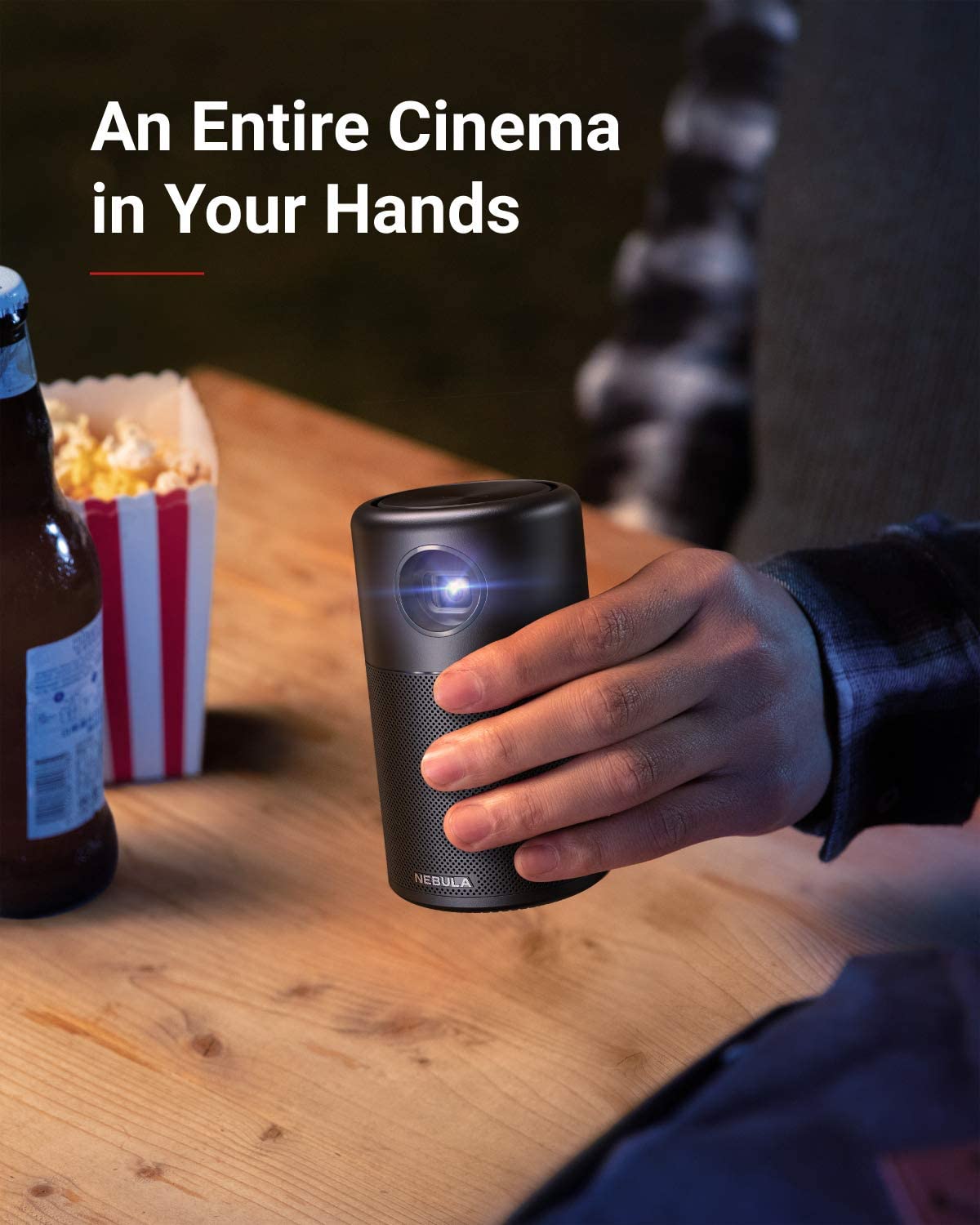 A hand holds a Nebula Capsule portable projector above a table with a bag of popcorn and a bottle of beer.